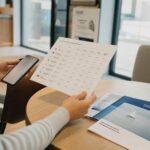 Osoba porównuje oferty funduszy na smartfonie i kartce przy stoliku w nowoczesnym wnętrzu.