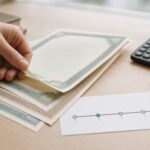 Dłoń przesuwa dokumenty obligacji na stole obok kalkulatora i długopisu.